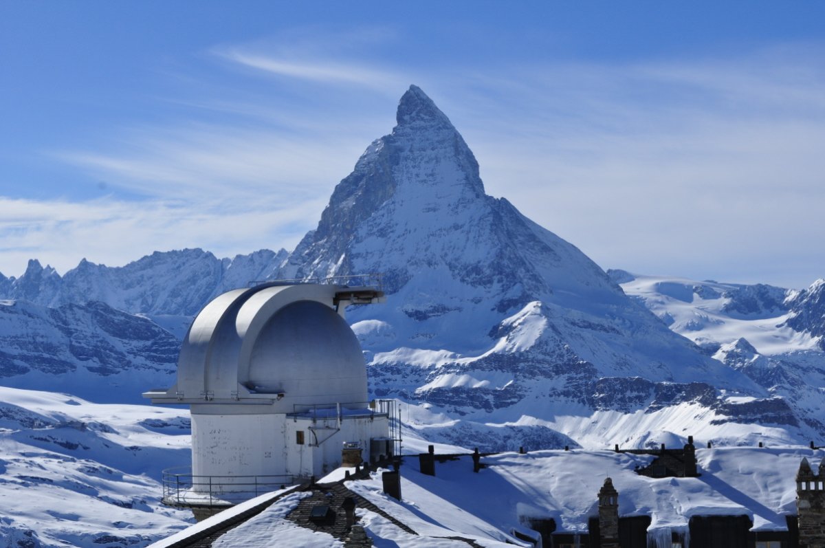 Im Vordergrund ist eine Kuppel einer Sternwarte zu sehen. In der Bildmitte ragt das Dreieck des Matterhorns in den Himmel. Links und rechts hat es kleinere Gipfel. Alle sind schneebedeckt.
