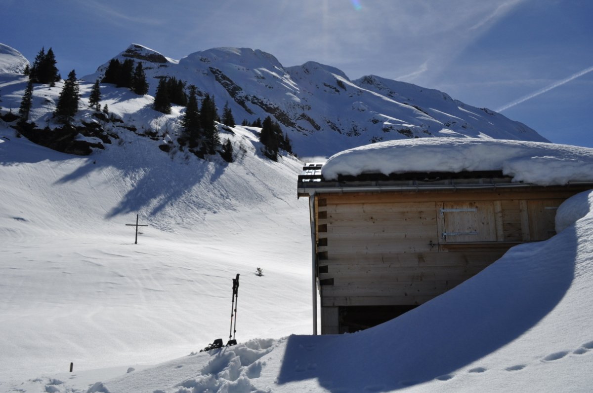 Rechts ragt ein Teil einer Alphütte aus dem Schnee. Im Hintergrund links ist ein Schneefeld, aus dem das Alpkreuz ragt. Dahinter ist ein Schneeabhang, der ober lockeren Wald trägt. Im Hintergrund ist die schneebedeckte Wasserberg-First zu sehen