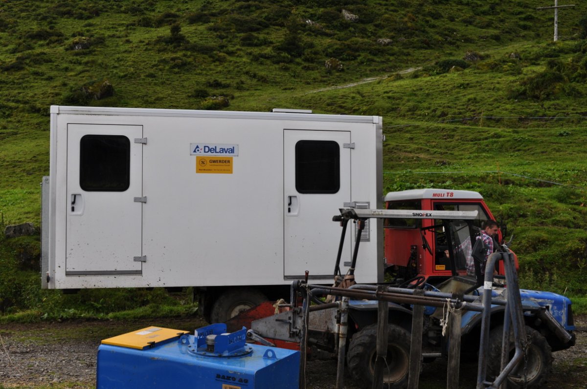 Auf der Alpstrasse steht der Transporter mit dem aufgeladenen Container. Im Vordergrund steht das Traktörli mit Heckschaufel, ein Schuänerstand und ein Dieseltank.