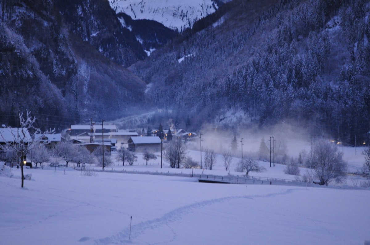 Im Vordergrund ist eine Wiese mit fast unberührtem Schnee. Dahinter ist eine Strasse, dann Wiese mit Bäumen, dann ein Fluss aus dem Nebel steigt. Links im Hintergrund sind Fabrikgebäude und ein Feriendorf zu erkennen. Darüber schliesst das Bild mit rauhreifüberzogenen Tannen an einem Abhang das Bild ab.