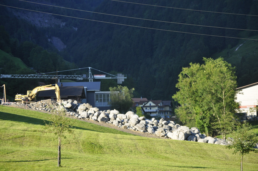 im Vordergrund eine abgemähte Wiese, quer durch das Bild ein langgezogener Haufen grosse Steine, die am Ufer des Teufbaches liegen, links ein Bagger, im Hintergrund Gebäude vom Viertel Stalden