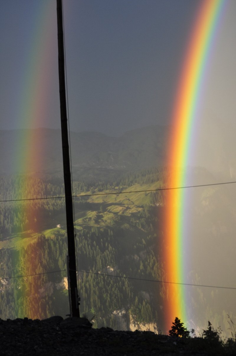 Ein Doppelregenbogen in kräftigen Farben, im Hintergrund die gegenüberliegende Alp mit Wald rundum.