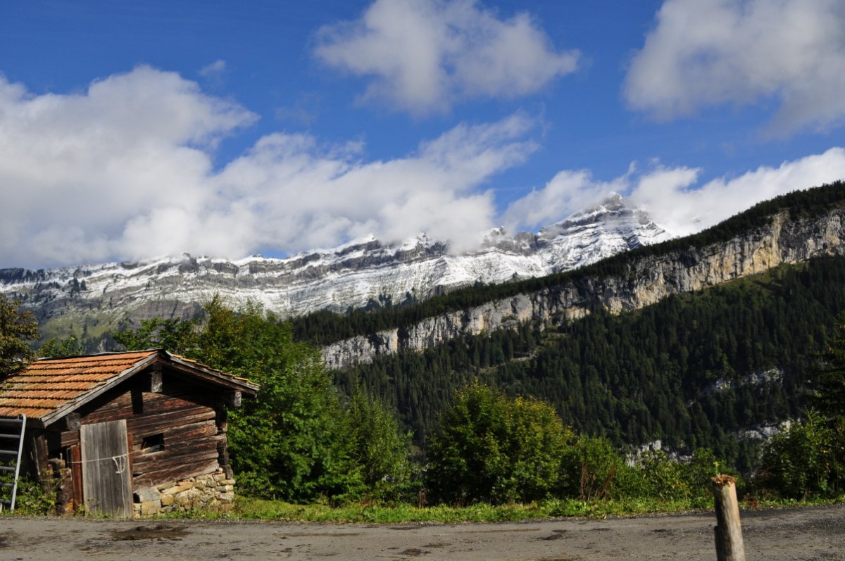 Links im Vordergrund eine alte kleine Alphütte, rechts davon ein Berg mit Felsband, im Hintergrund der Heubrig weiss überschneit,darüber ein blauer Himmel mit Wolkenfetzen.