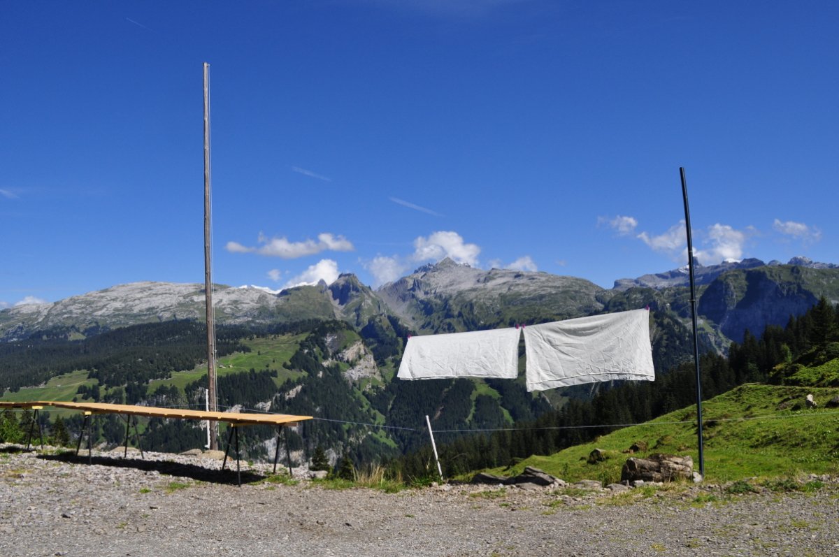 Im Vordergrund ist ein Schotterplatz. Darauf ist ein Gestell aufgestellt, auf dem gewaschenen Käsebretter trocknen. Am Rand des Platzes ist zwischen zwei hohen Pfosten ein dünnes Drahtseil gespannt. Daran hängen zwei Blächen (Käsetücher) zum Trocknen. Im Hintergrund ist ein Panorama mit Alpweiden, Wald und Karren in Grau. Darüber wölbt sich ein 'chnütschblauer' Himmel.
